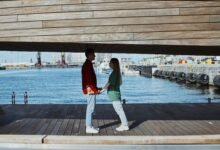 A couple holds hands on a wooden bridge overlooking a city waterfront, capturing a romantic moment.