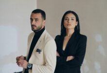 A man and woman posing confidently in stylish formal wear against a textured wall.