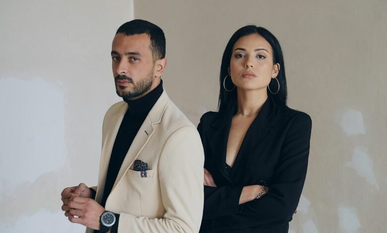 A man and woman posing confidently in stylish formal wear against a textured wall.